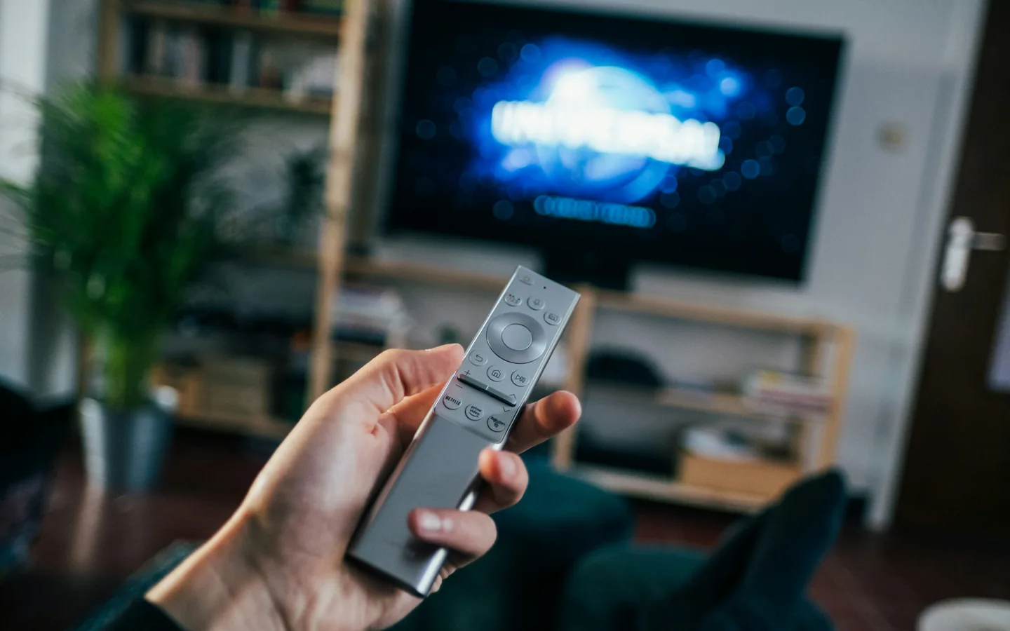 As perguntas certas antes de pagar economizam meses de arrependimento. Mão segurando um controle remoto prateado apontado para um televisor desfocado ao fundo