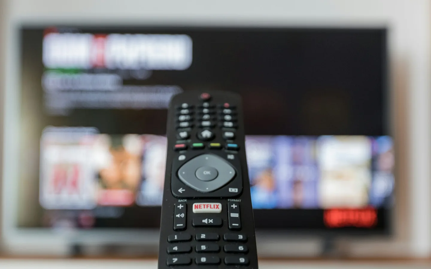 Perfis e bloqueio por senha são configurados dentro do próprio aplicativo. Controle remoto preto em primeiro plano diante de um televisor desfocado exibindo um catálogo de títulos